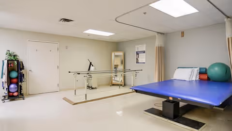 A physical therapy or rehabilitation room with a blue therapy table, exercise balls, parallel bars for walking practice, a mirror, and a small rack holding various exercise equipment against a beige wall.