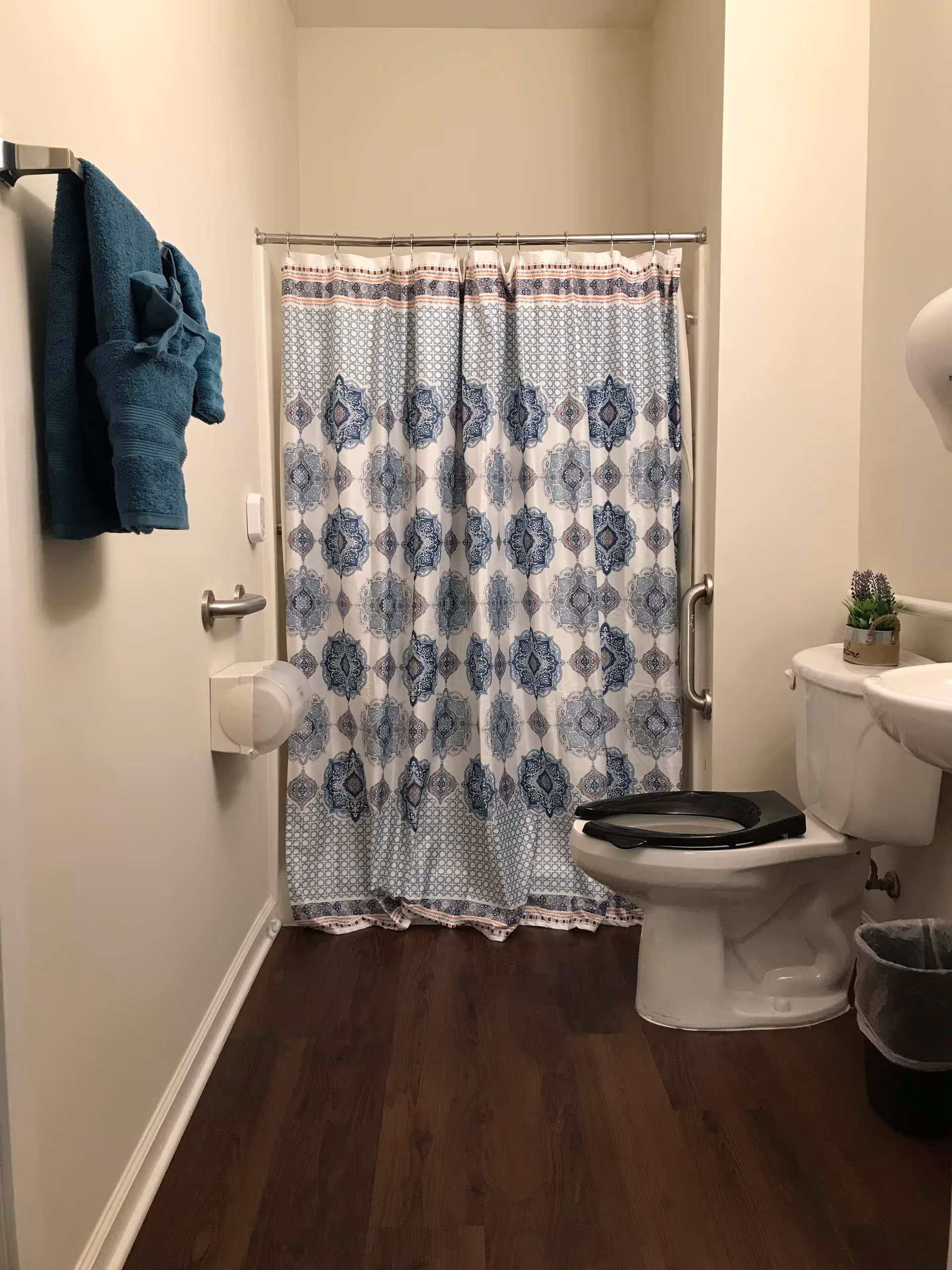 Bathroom showing a patterned blue-and-white shower curtain, toilet with a raised seat, sink, and teal towels on a wall rack.