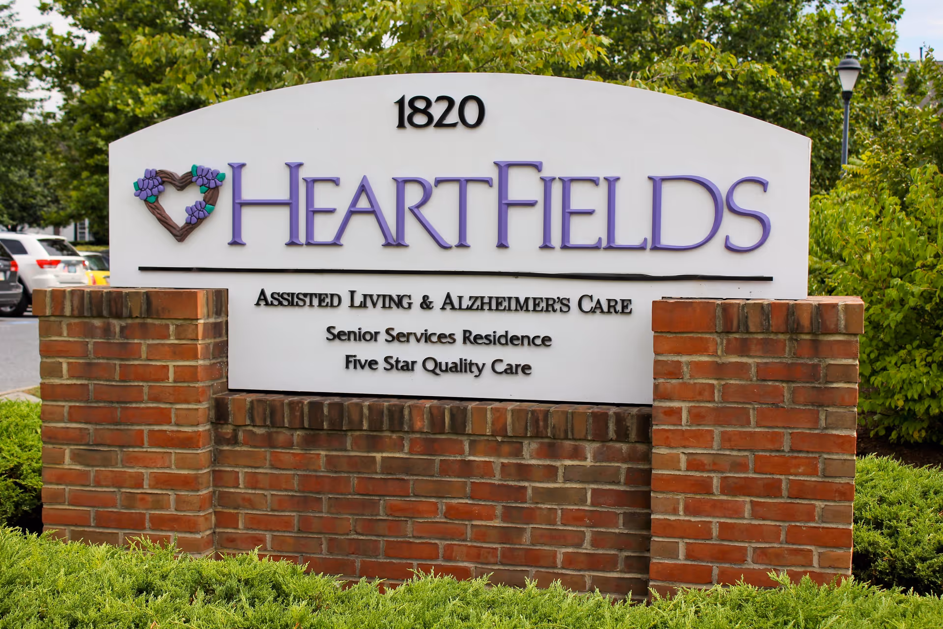 A large outdoor sign for HeartFields Assisted Living & Alzheimer's Care, a senior services residence offering five star quality care. The sign is mounted on a brick base with greenery around it and trees and parked cars visible in the background.
