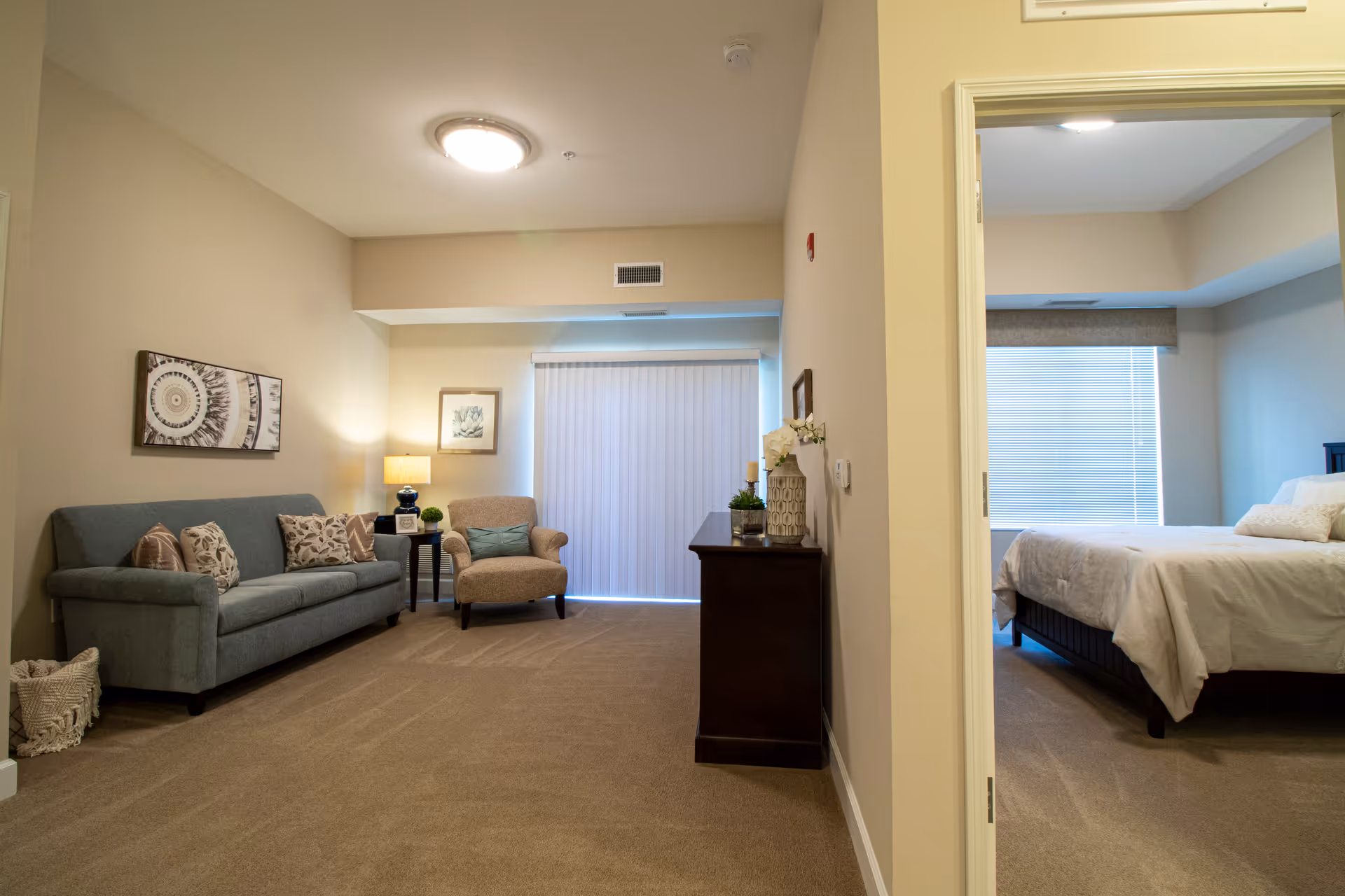 A cozy living room area with a blue sofa adorned with patterned pillows, a beige armchair with a blue cushion, a side table with a lamp, and framed artwork on the walls. To the right, an open doorway reveals a bedroom with a neatly made bed and a window with blinds.