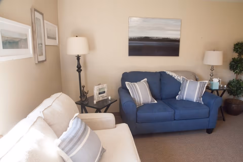 A cozy living room with a beige sofa and a blue loveseat, each adorned with striped throw pillows. There are two side tables with lamps, a small decorative statue, a picture frame, and a tissue box. The walls are beige with framed artwork and a large abstract painting above the loveseat. A potted plant is in the corner.