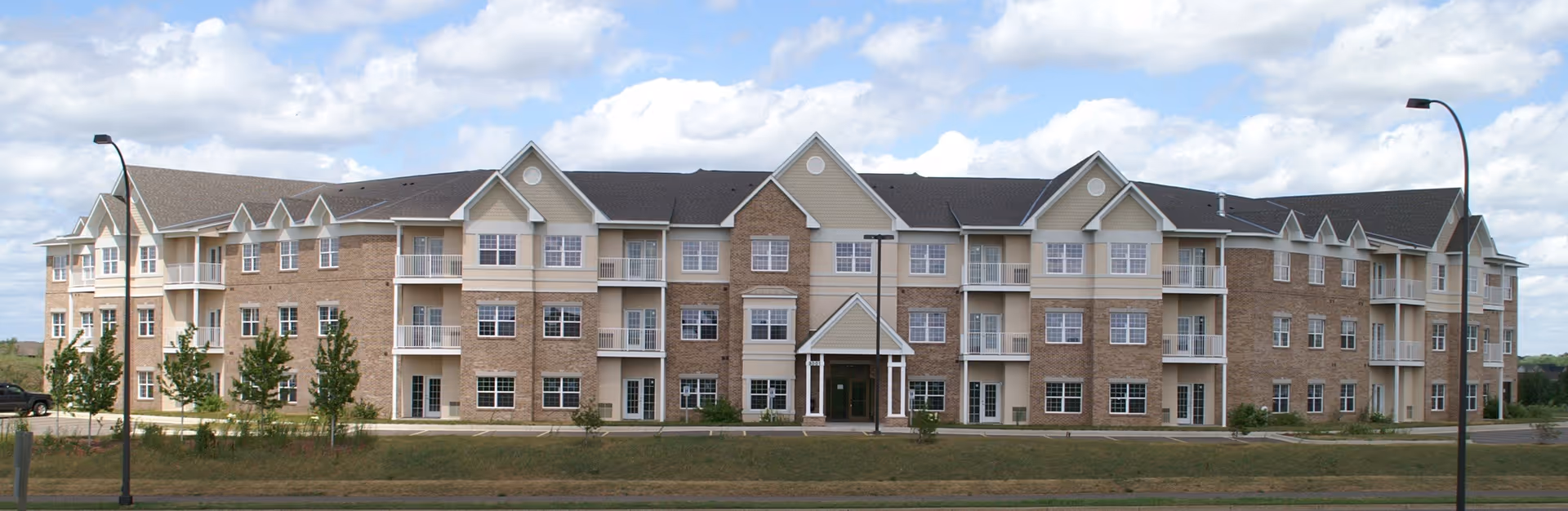 Exterior front view of a large three-story senior living facility building with beige and brick facade, multiple windows, balconies, and a central entrance under a peaked roof. The building is surrounded by a grassy area with small trees and street lamps under a partly cloudy sky.