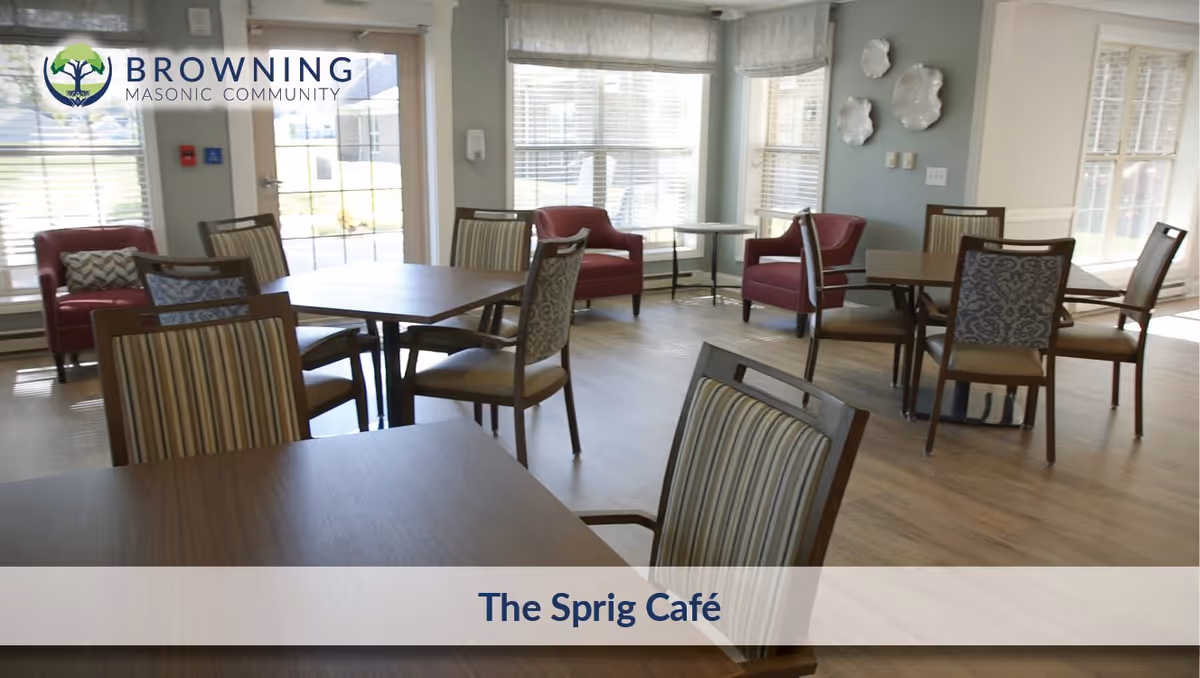 A bright and airy café area with several wooden tables and chairs arranged neatly. The room has large windows letting in natural light, with red armchairs placed near the windows. The walls are painted light gray with decorative plates hanging on one wall. The floor is wooden, and there is a door leading outside. The image includes the text 'Browning Masonic Community' and 'The Sprig Café'.