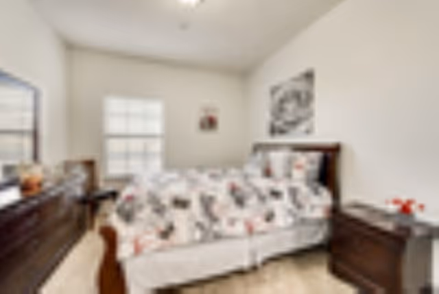 A bedroom with a bed featuring a patterned comforter and pillows, a wooden dresser with a mirror, a nightstand with a small decorative item, and two windows letting in natural light.