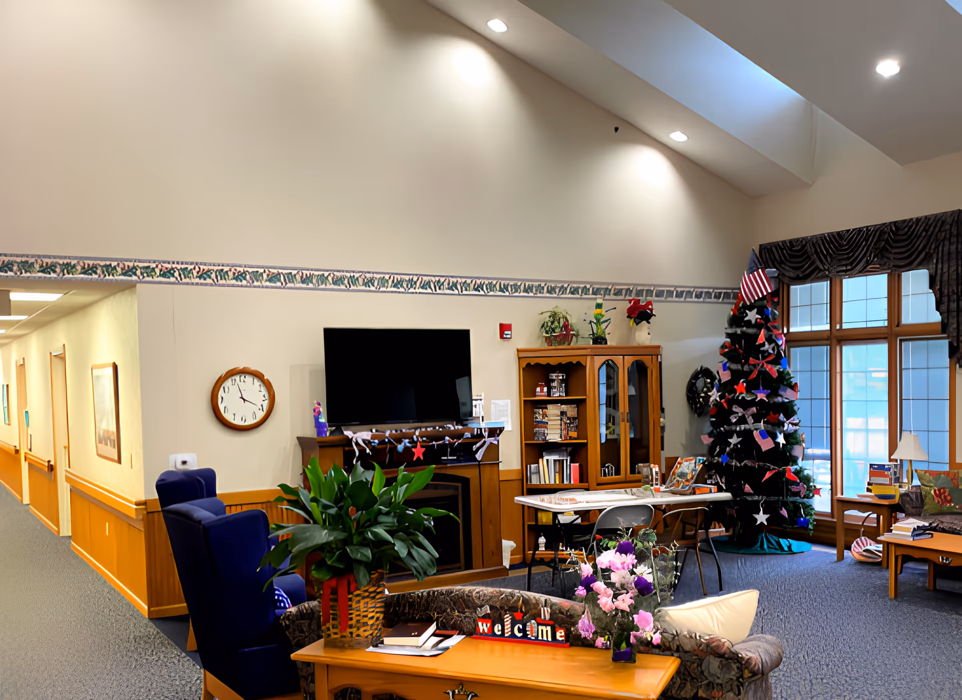 A cozy living room area in Azria Health Rose Vista featuring a large flat-screen TV mounted above a fireplace, a decorated Christmas tree with an American flag on top, a wooden cabinet with books and decorations, a table with chairs, and comfortable seating including a blue armchair and a floral-patterned couch. The room has high ceilings with recessed lighting and large windows with dark curtains letting in natural light. A hallway is visible to the left with framed pictures on the walls.