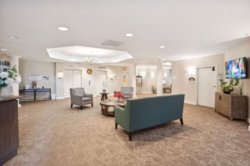 Spacious senior living facility lobby with seating area, coffee table, elevators and a wall-mounted TV.