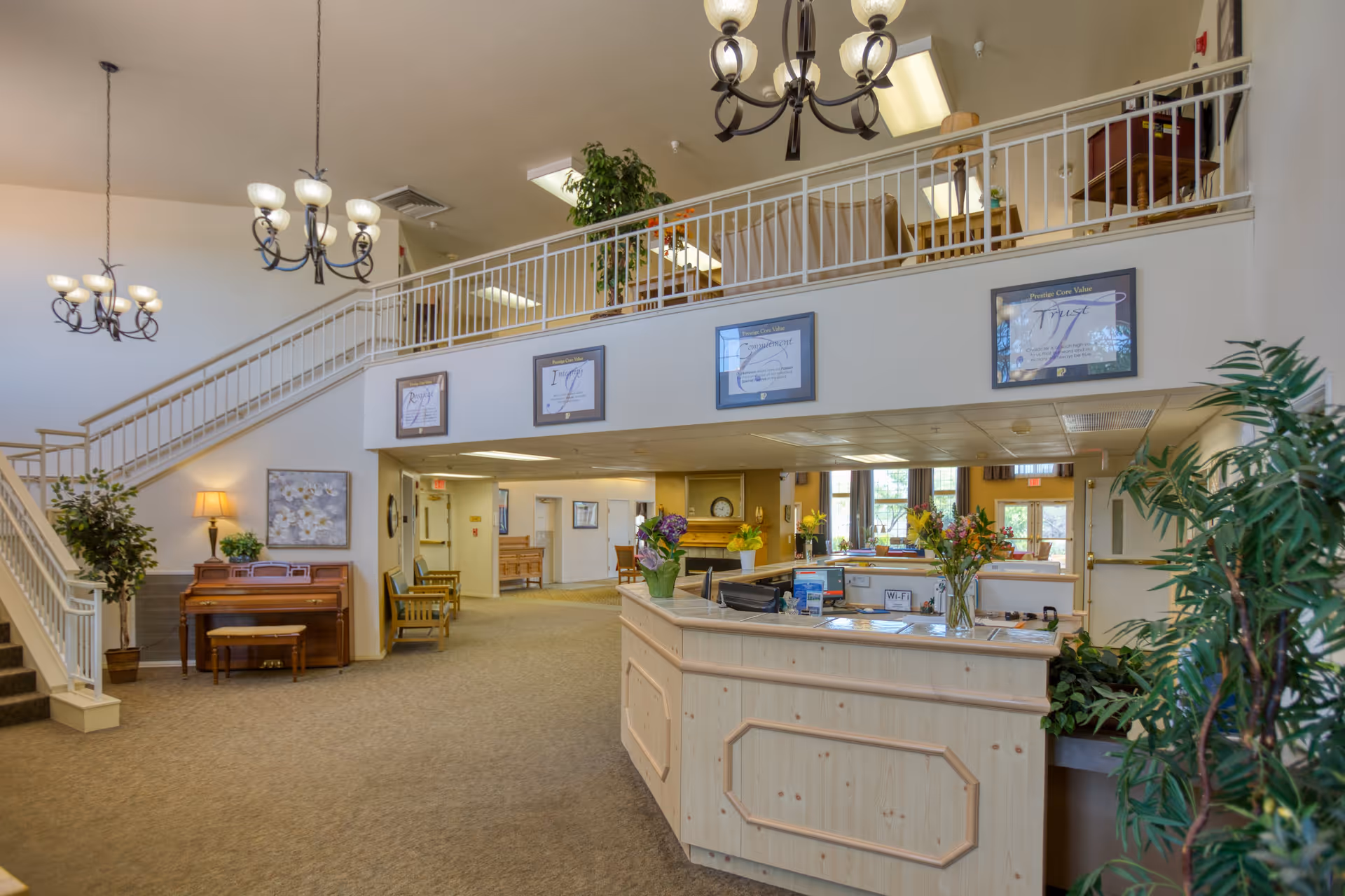 Interior view of a senior living facility reception area with a wooden front desk adorned with flowers and informational materials. Behind the desk is a seating area with large windows letting in natural light. To the left, there is a staircase leading to an upper level with a railing, and a piano with a bench and a lamp beside it. The space is decorated with framed pictures, plants, and hanging chandeliers providing warm lighting.