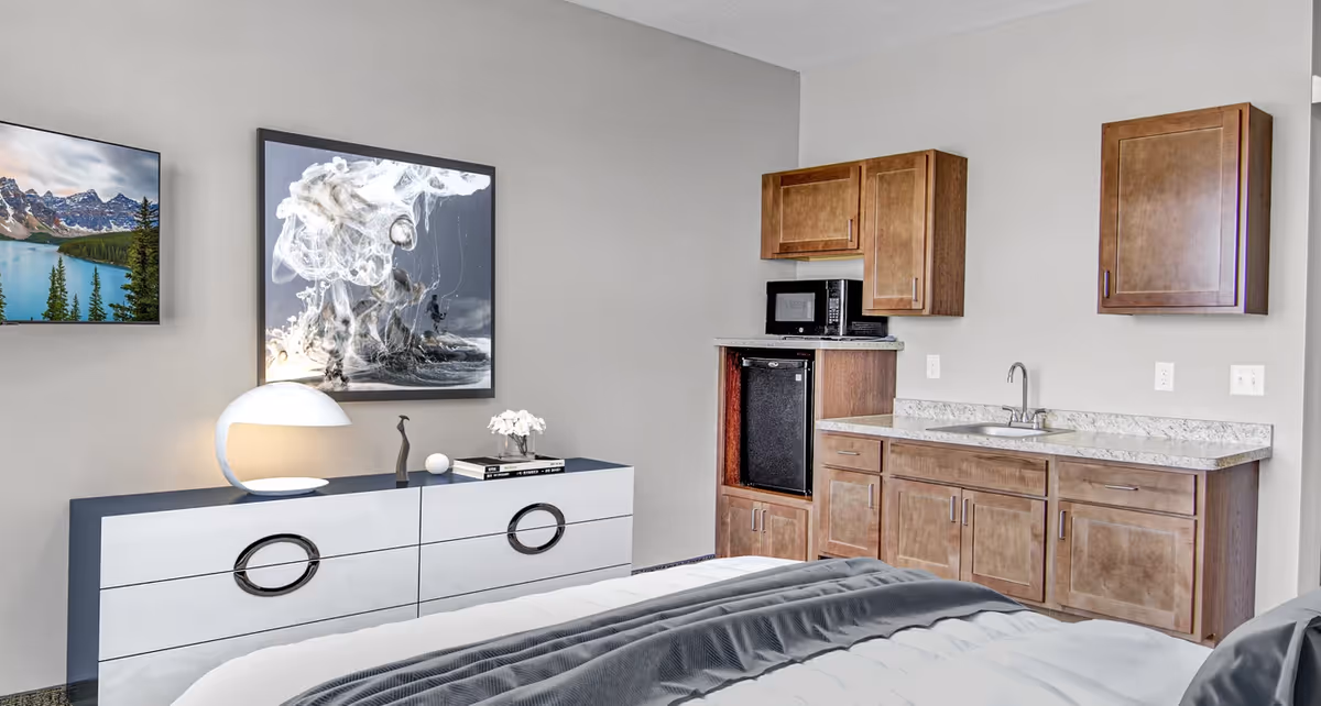 Studio-style bedroom suite with a bed in the foreground, a dresser and artwork on the wall, and a kitchenette area with cabinets, sink, microwave and mini-fridge.
