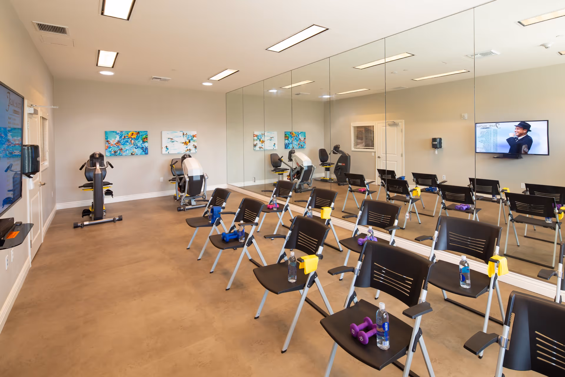 A fitness room with several black chairs arranged in rows, each chair holding a water bottle, small dumbbells, and a yellow towel. The room has exercise equipment against the far wall, large mirrors covering one side, colorful artwork on the walls, and a TV mounted on the wall displaying a person in a hat.