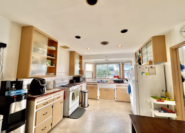 A bright kitchen with beige cabinets and white appliances including a stove and refrigerator. There is a large window above the sink letting in natural light. Various kitchen items such as a microwave, coffee maker, and dish rack are visible on the countertops. The floor is tiled and the ceiling has recessed lighting.