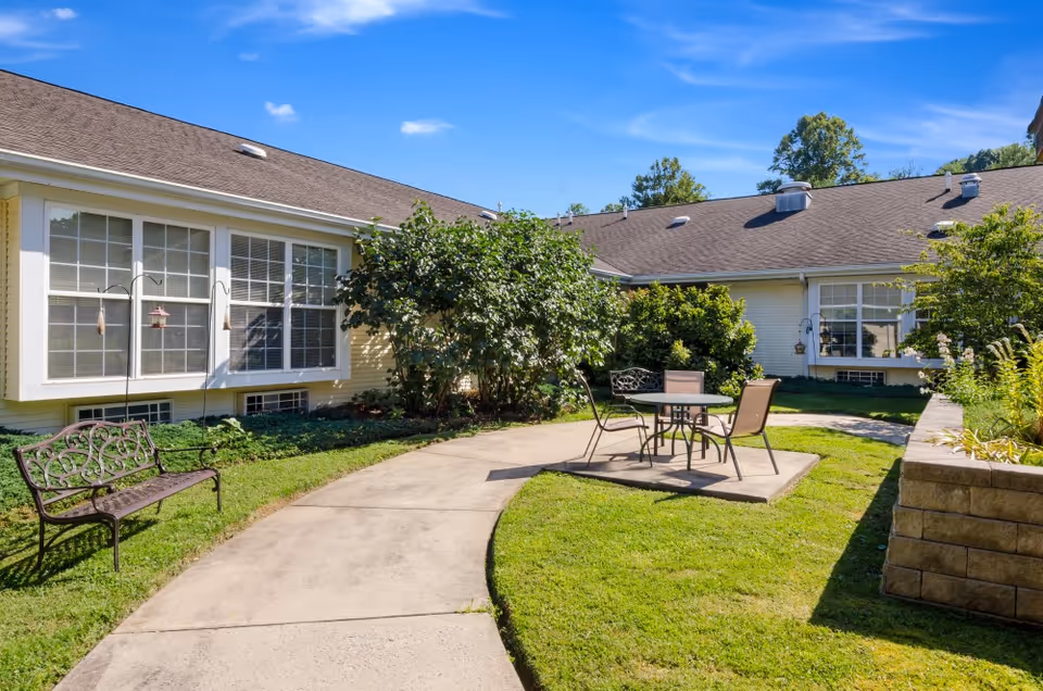 Outdoor courtyard area at American House Bristol featuring a curved concrete walkway, a metal bench, a small table with two chairs on a concrete pad, surrounded by green grass, bushes, and trees under a clear blue sky.