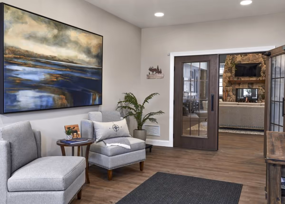 A cozy interior seating area with two gray upholstered chairs and a small round wooden table between them. A large abstract painting with blue and brown tones hangs on the wall above the chairs. A potted plant is placed near the corner. In the background, there is a wooden sliding door with glass panels leading to another room featuring a stone fireplace with a mounted TV above it and a beige sofa.