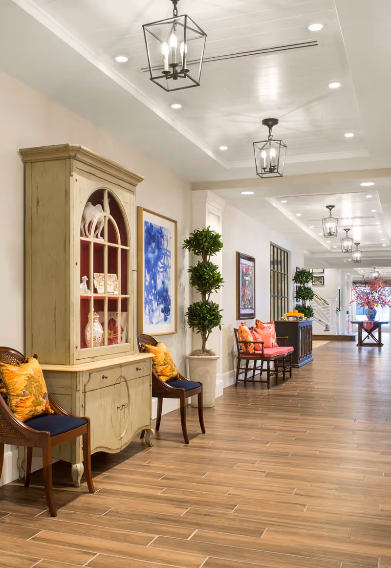 A bright and spacious hallway in a senior living facility with wooden flooring, decorative chairs with cushions, a vintage cabinet displaying ceramic items, framed artwork on the walls, potted plants, and modern ceiling lights.