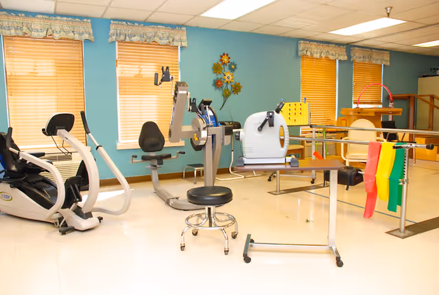 A brightly lit rehabilitation room with exercise machines, parallel bars, colorful resistance bands, and a rolling stool in front of teal walls and windows.