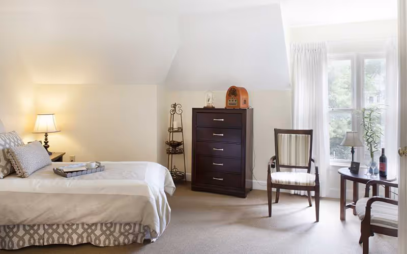 A bright bedroom with a single bed covered in white and patterned bedding, a bedside table with a lamp, a dark wooden chest of drawers, a decorative metal stand, two wooden chairs with striped cushions, and a small round table with a lamp, wine bottle, and glass near a window with white curtains.