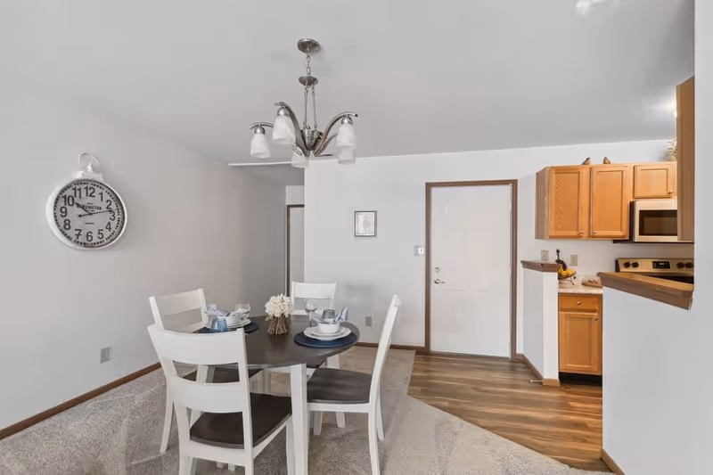 Interior view of a dining area with a round table set for four with white chairs. A decorative wall clock is mounted on the left wall. The dining area has carpet flooring, and adjacent to it is a kitchen with wooden cabinets, a microwave, and a stove. The kitchen area has wood flooring, and a white door is visible in the background.