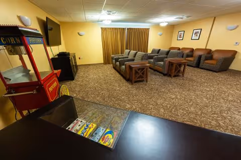 A cozy common area with several cushioned armchairs and two wooden side tables arranged on a carpeted floor. The walls are painted light yellow and decorated with framed pictures. A popcorn machine is visible in the foreground, along with a counter displaying various snacks. The room is softly lit with ceiling lights and wall sconces, and a large window is covered by beige curtains.