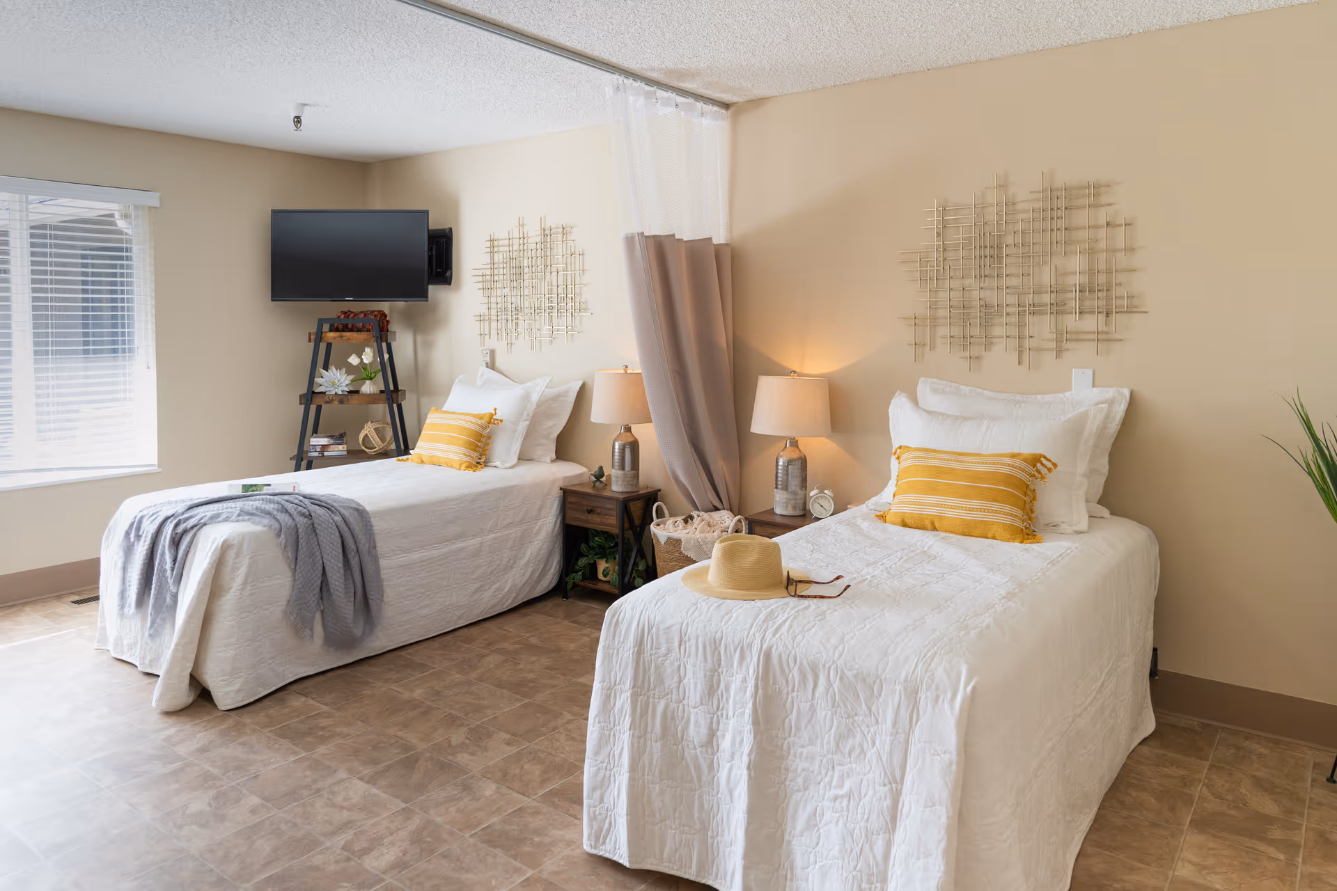 A bright and clean room with two single beds, each made with white bedding and yellow striped pillows. Between the beds is a small wooden nightstand with two lamps and a basket underneath. A flat-screen TV is mounted on the wall above a small shelf unit holding decorative items. The room has beige walls, a window with blinds, and a curtain hanging from a ceiling track between the beds.