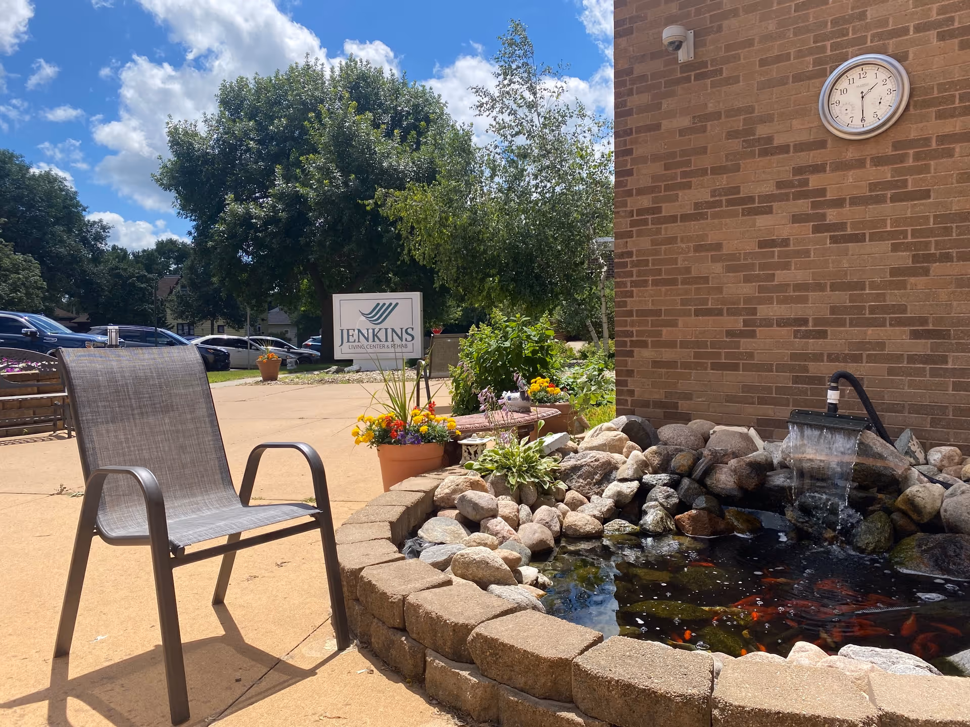 Outdoor patio area at Jenkin's Living Center & Rehab featuring a single chair, a small pond with rocks and a water fountain, colorful flower pots, a brick wall with a clock, and a sign with the facility's name in the background under a partly cloudy blue sky.