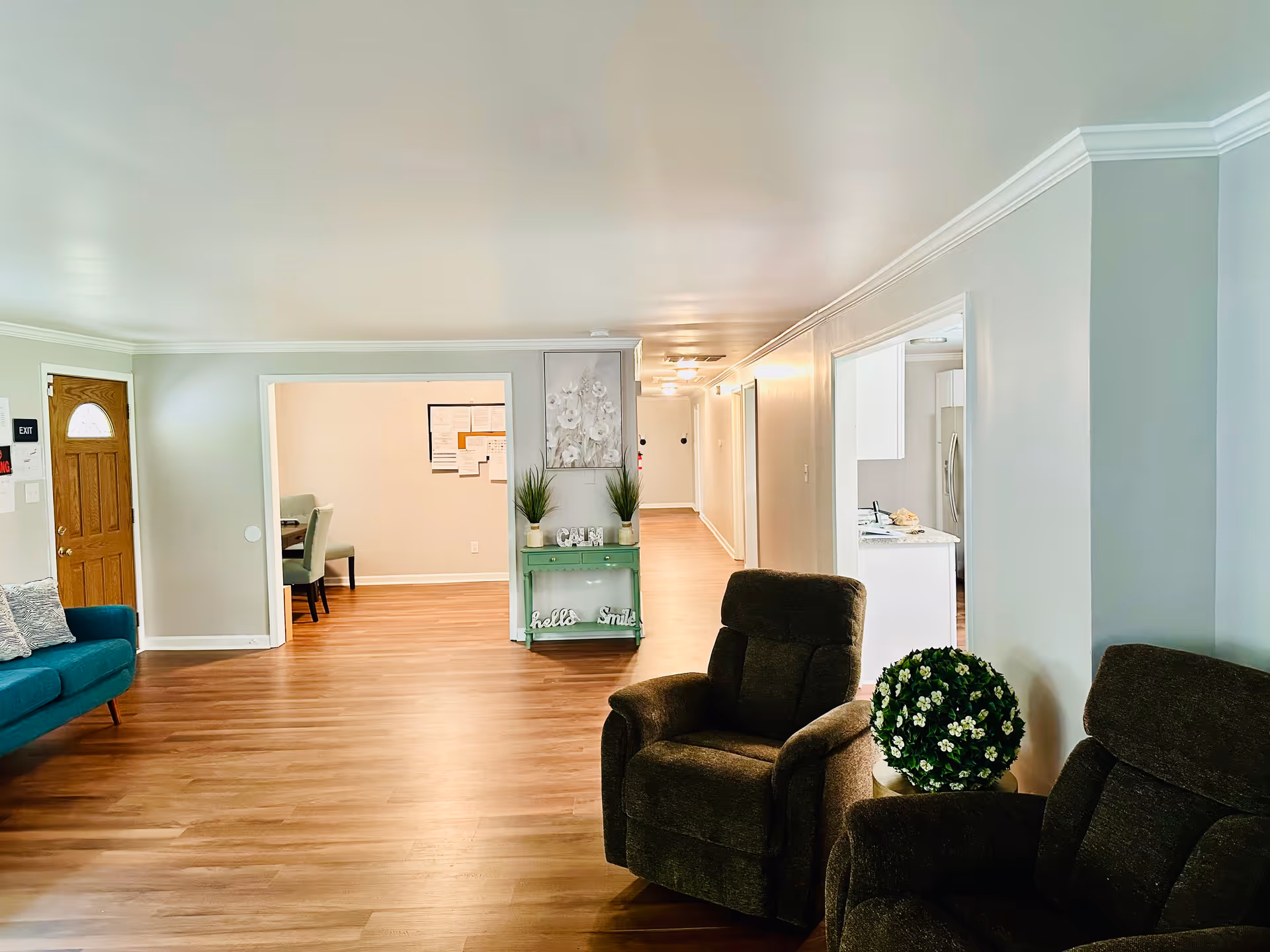 A bright and spacious living room area in an assisted living facility with wooden flooring, a teal sofa with cushions on the left, two dark brown recliner chairs on the right, and a green console table with plants and decorative items in the center. There is a wooden door with a small window on the left wall and an open doorway leading to a dining area with chairs. The walls are painted light gray, and the ceiling is white with crown molding.