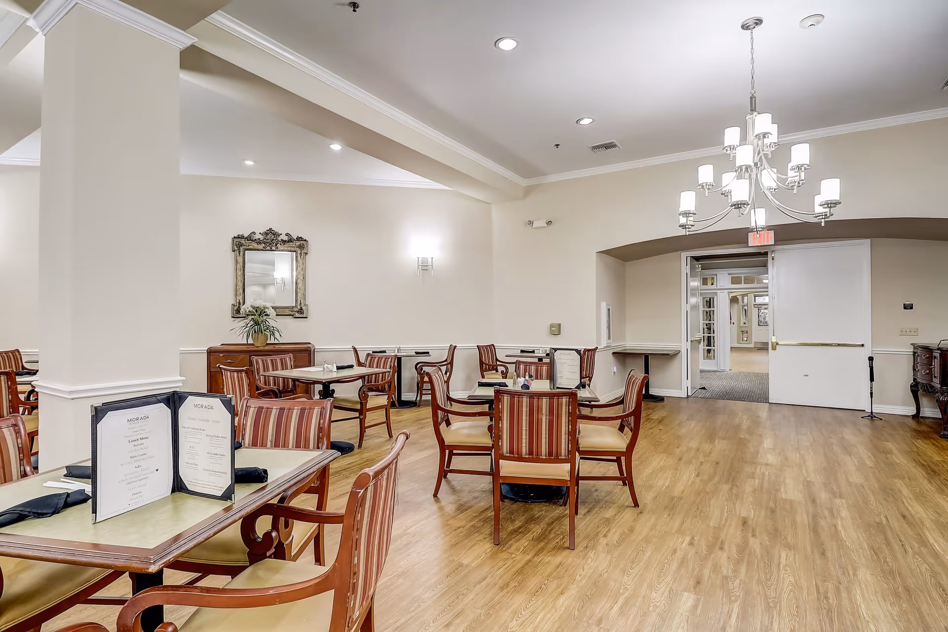 A dining room in Morada Cedar Hill featuring several tables with striped cushioned chairs. Menus and black napkins are placed on the tables. The room has light-colored walls, wood flooring, a chandelier, and a decorative mirror on the wall. There is an open doorway leading to another area.