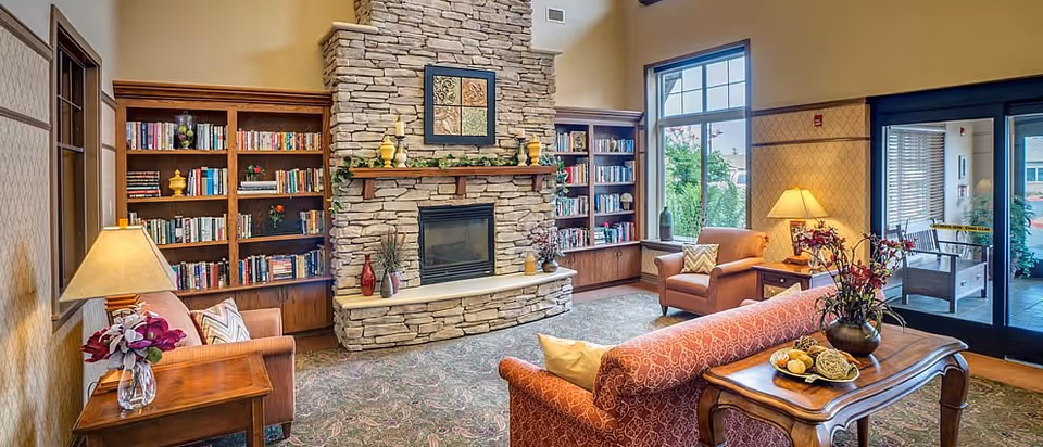 A cozy living room area with a stone fireplace in the center, flanked by wooden bookshelves filled with books. The room has comfortable armchairs and a sofa with decorative pillows, a wooden side table with a lamp and flowers, and a coffee table with a floral arrangement and decorative items. Large windows and glass doors let in natural light and show some greenery outside.