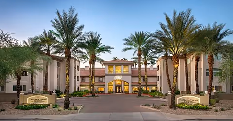 Front exterior view of The Ranch Estates At Scottsdale facility with a driveway entrance lined by tall palm trees and two signs reading 'The Manor Village'. The building has a central entrance with large windows and a tiled roof under a clear blue sky.