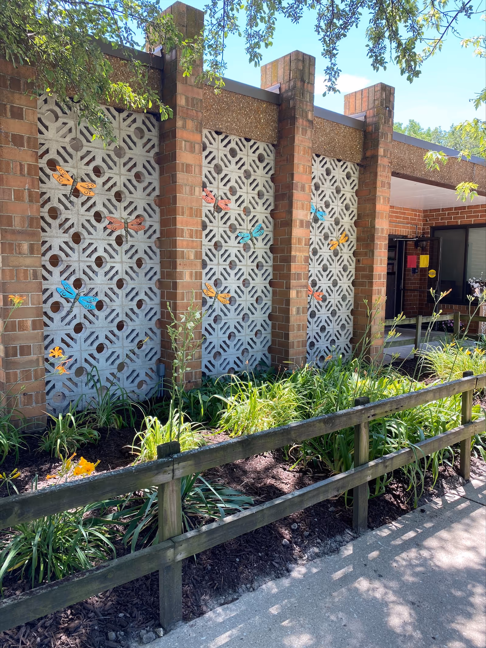Exterior view of a brick building with decorative concrete block panels adorned with colorful butterfly decorations. There is a small garden bed with green plants and flowers in front, bordered by a wooden fence. The entrance to the building is visible on the right side under a covered area.