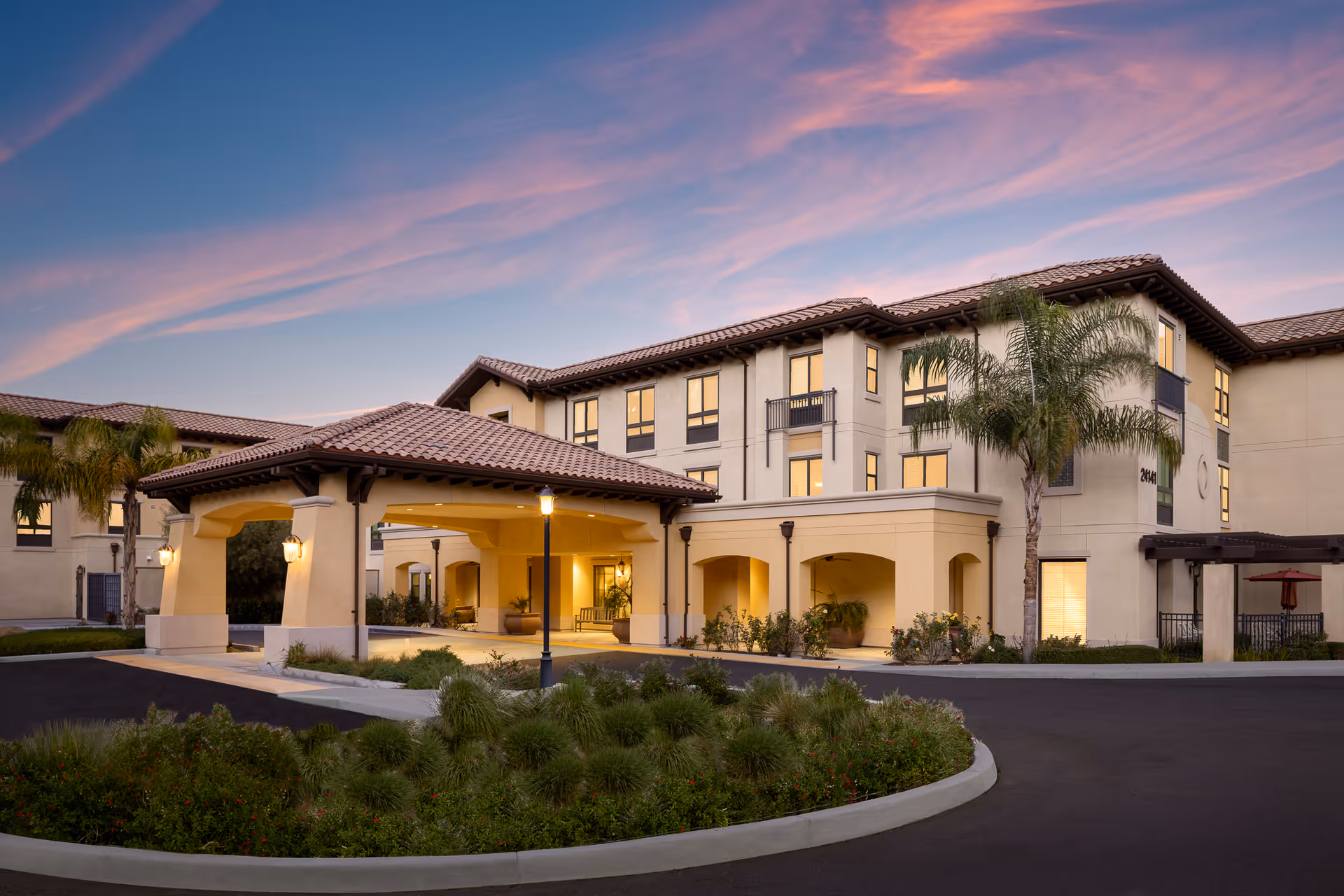 Exterior view of Belmont Village Senior Living Calabasas building at dusk with a covered entrance, palm trees, and landscaped greenery under a colorful sky.