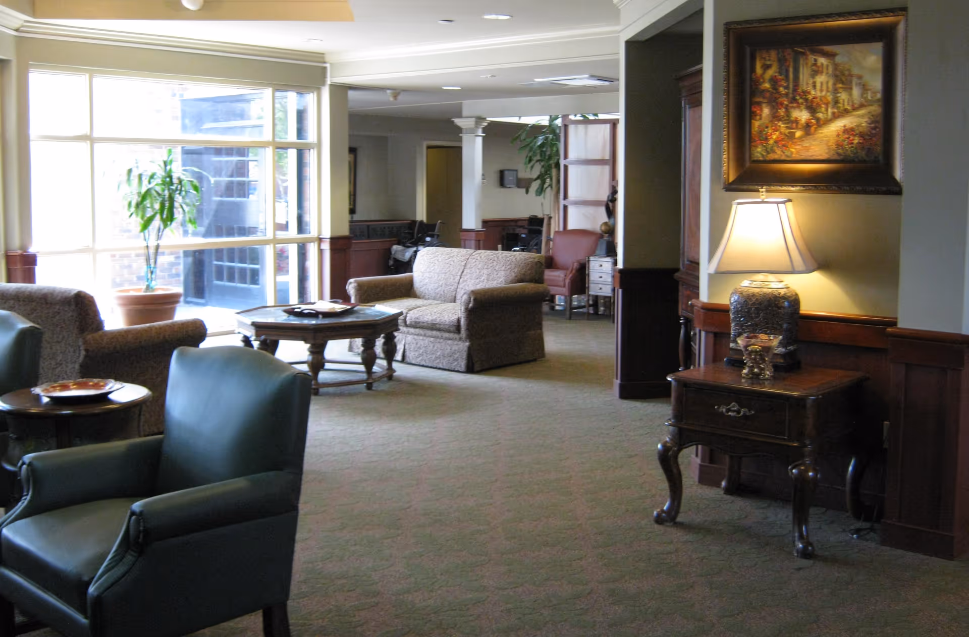 A well-lit living room area in a senior living facility with multiple upholstered chairs and sofas arranged around wooden coffee tables. There is a large window letting in natural light, a potted plant near the window, and a side table with a decorative lamp and a framed painting on the wall.
