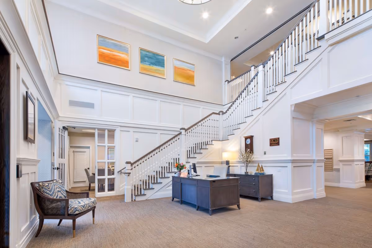 A spacious and well-lit interior area of a senior living facility featuring a large staircase with white railings leading to an upper floor. There is a dark wooden reception desk with a computer and decorative items, a matching sideboard with a clock and vase, and a patterned armchair. The walls are white with paneling, and three colorful abstract paintings hang above the staircase. The carpet is beige, and the ceiling has recessed lighting.