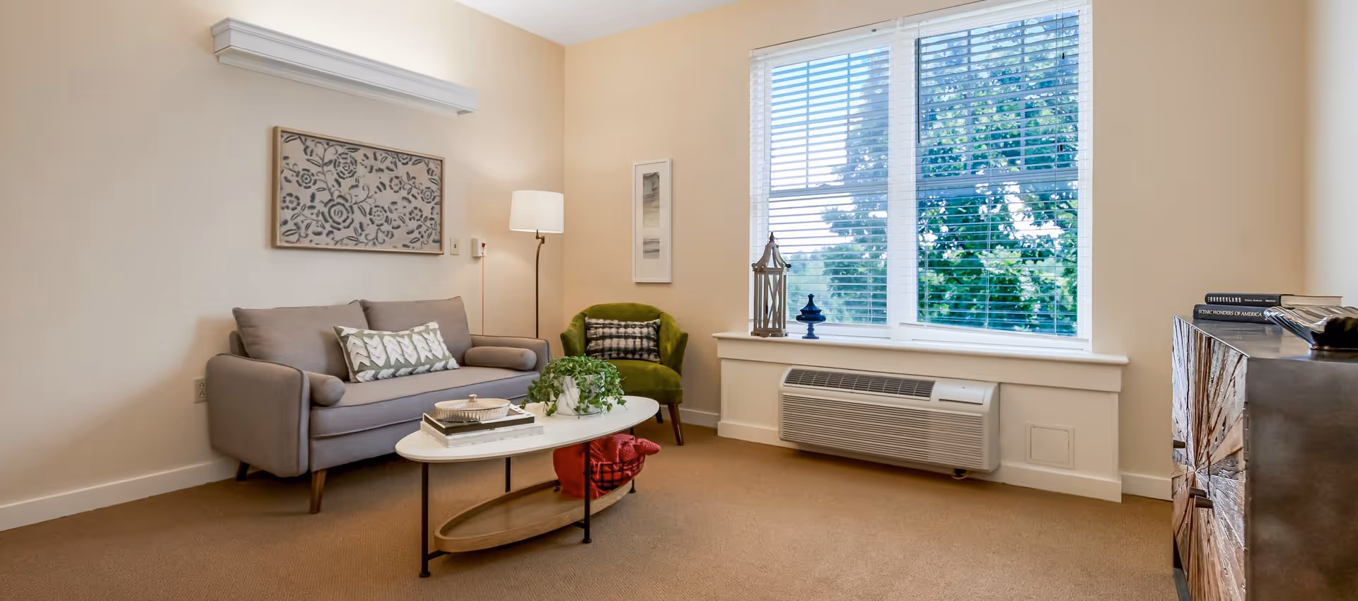 A cozy sitting area with a gray sofa, green armchair, coffee table, wall art, and a large window with blinds.