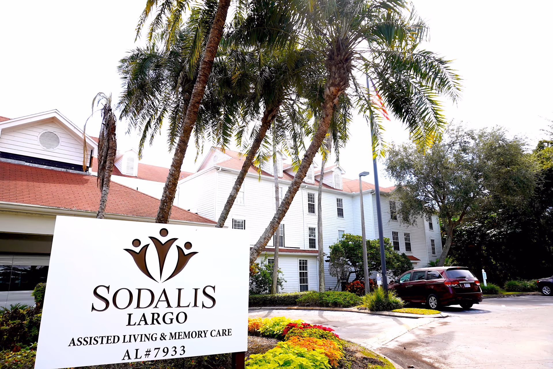 Exterior view of Sodalis Largo assisted living and memory care facility with a white multi-story building, palm trees, a parking lot with cars, and a large sign in the foreground displaying the facility name and license number.