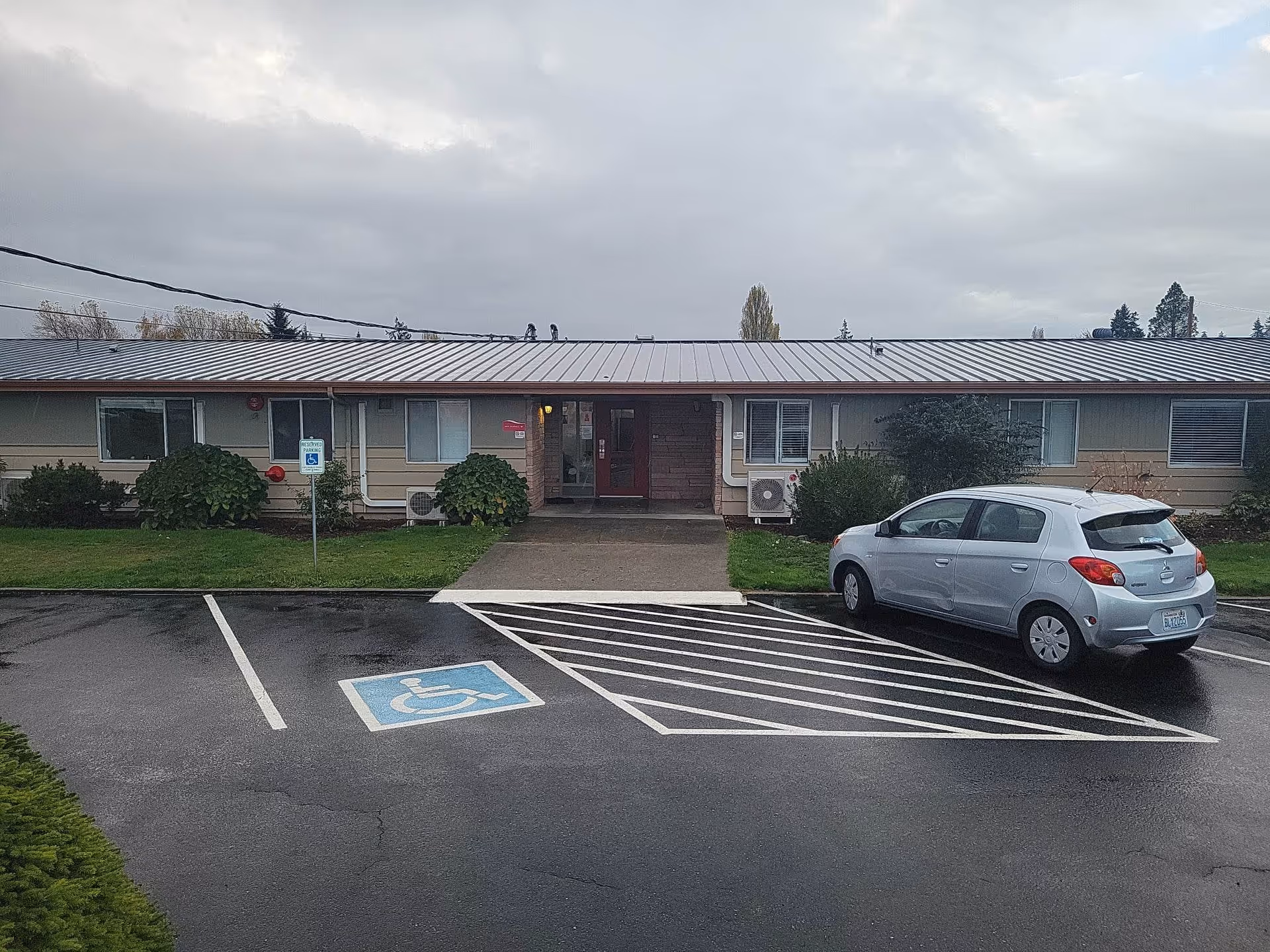 Exterior view of a single-story building with a metal roof and a central entrance. There is a parking lot in front with a reserved handicapped parking space marked in blue and white, and a silver car parked next to it. The sky is overcast and there are some bushes and grass around the building.