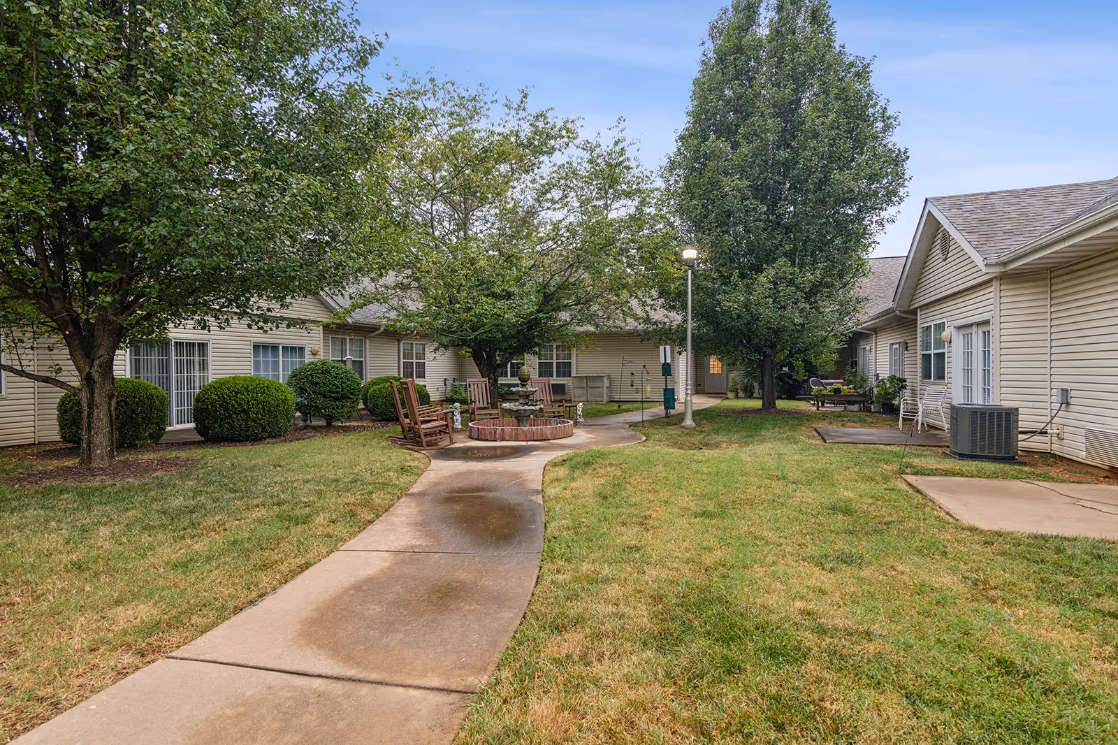 Outdoor courtyard area at The Bungalows at Branson Meadows featuring a curved concrete walkway, green grass, several trees, bushes, wooden rocking chairs around a circular brick fire pit, and beige single-story buildings with white-framed windows and doors surrounding the space.