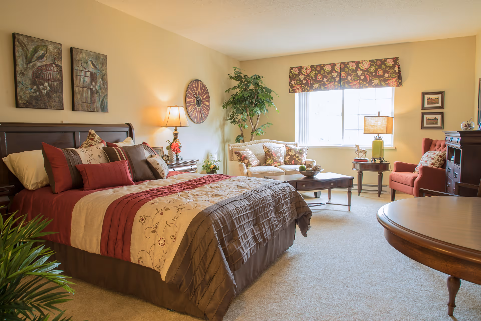 A well-decorated bedroom with a large bed featuring multiple pillows and a patterned bedspread. The room includes a nightstand with a lamp, a wall clock, two paintings of birds in cages, a potted plant, a beige loveseat with floral cushions, a wooden coffee table, a red armchair, and a window with floral valance allowing natural light to fill the room.