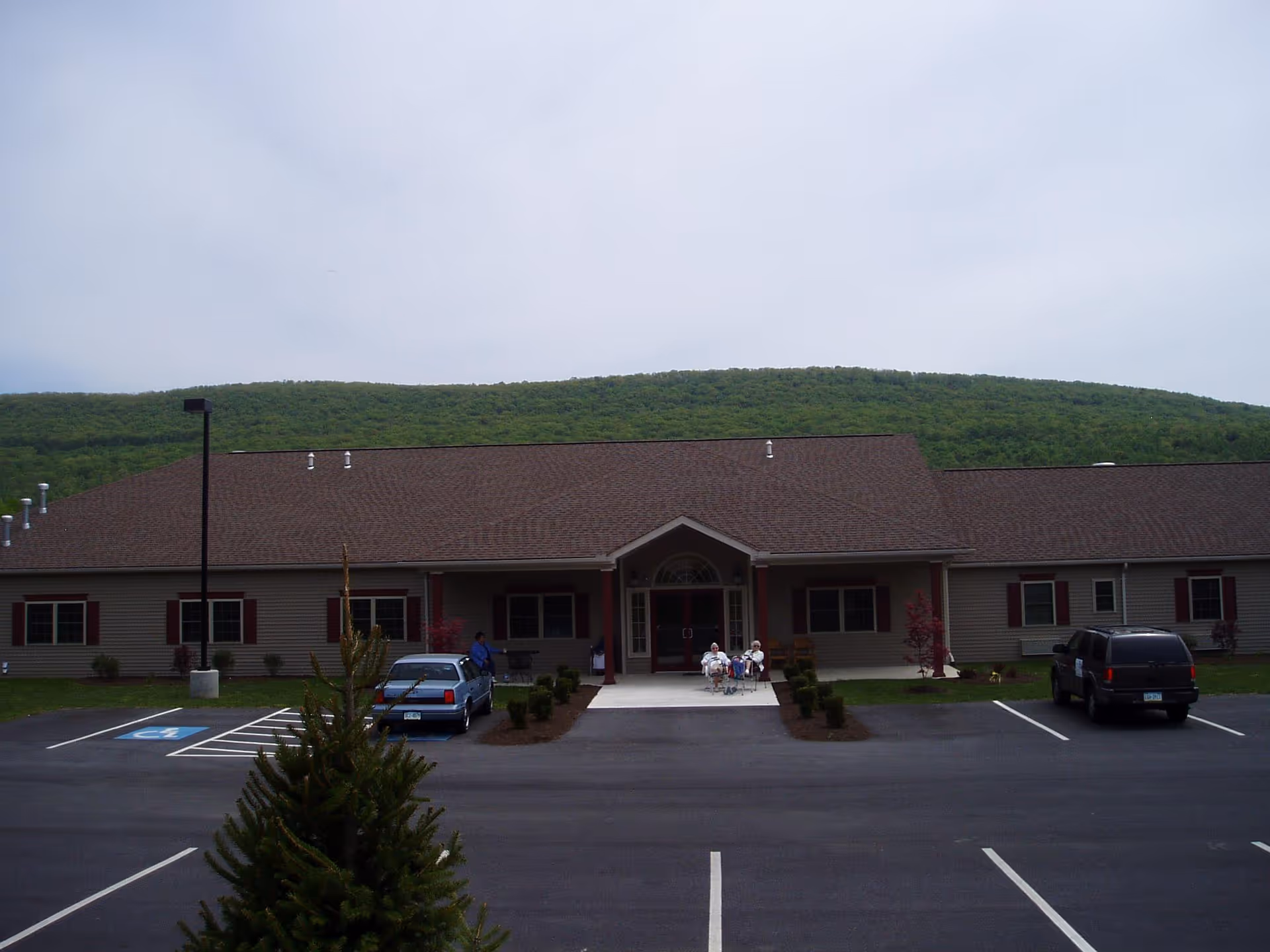Front exterior view of Olivia Village Assisted Living facility with a parking lot in front, a few cars parked, and a group of people sitting near the entrance. The building has a brown roof and beige siding with red window shutters. Green hills are visible in the background under a cloudy sky.