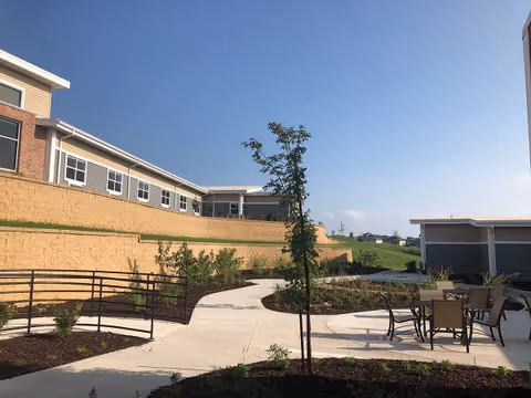 Sunny exterior view of Heritage Manor with a curved walkway, landscaping, and outdoor dining tables.