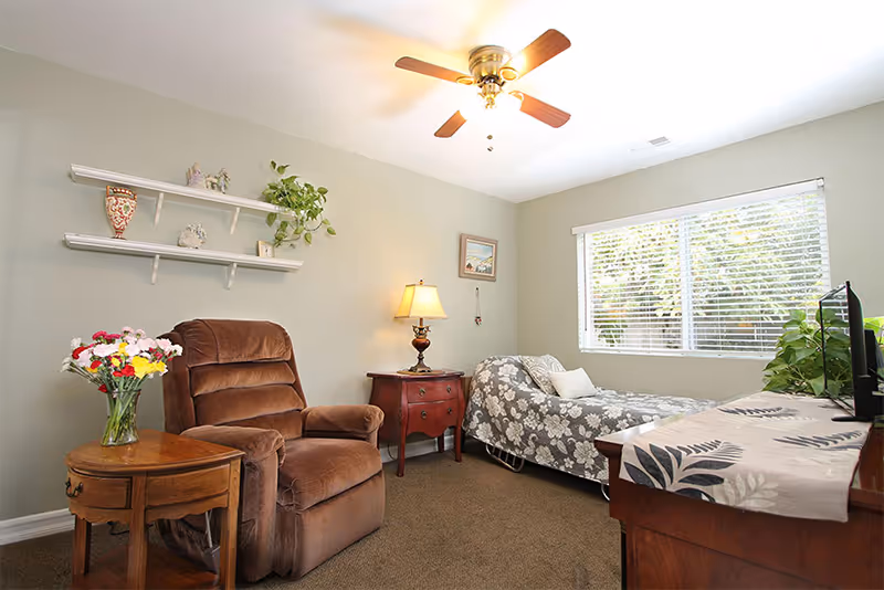 A cozy bedroom with a single bed covered in a floral patterned bedspread, a brown recliner chair, a wooden side table with a vase of colorful flowers, a wooden nightstand with a lamp, two white wall shelves with decorative items, a ceiling fan with wooden blades, and a large window with white blinds letting in natural light.