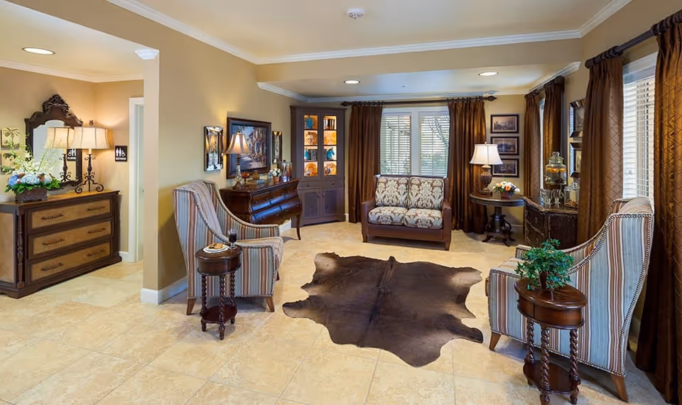 A cozy living room area with beige walls and tiled floors, featuring two striped armchairs with small round wooden side tables, a patterned loveseat, a dark wooden cabinet with glass doors, and a dark brown animal hide rug in the center. There are large windows with brown curtains, framed pictures on the walls, and decorative lamps providing warm lighting. A dresser with a lamp and a mirror is visible near an open doorway leading to another room.