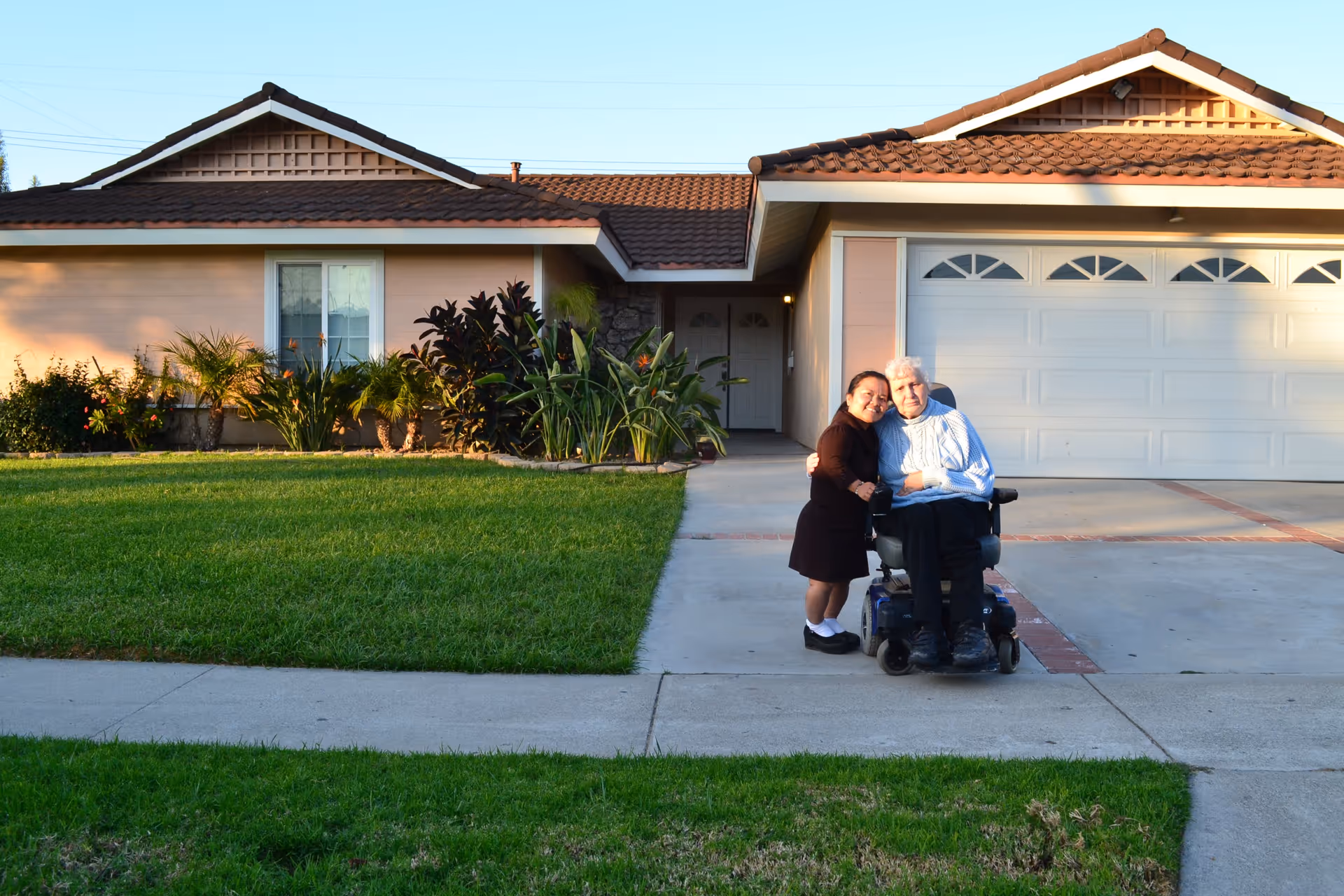 Two people, one standing and one in a wheelchair, embracing and smiling in front of a single-story house with a well-maintained lawn and plants near the entrance.