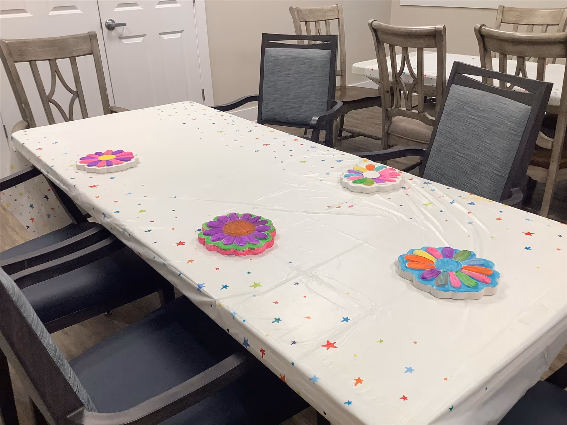 A rectangular table covered with a white tablecloth decorated with colorful stars and four colorful flower-shaped coasters. The table is surrounded by various chairs, some with armrests and some without. The room has light-colored walls and a closed white door in the background.