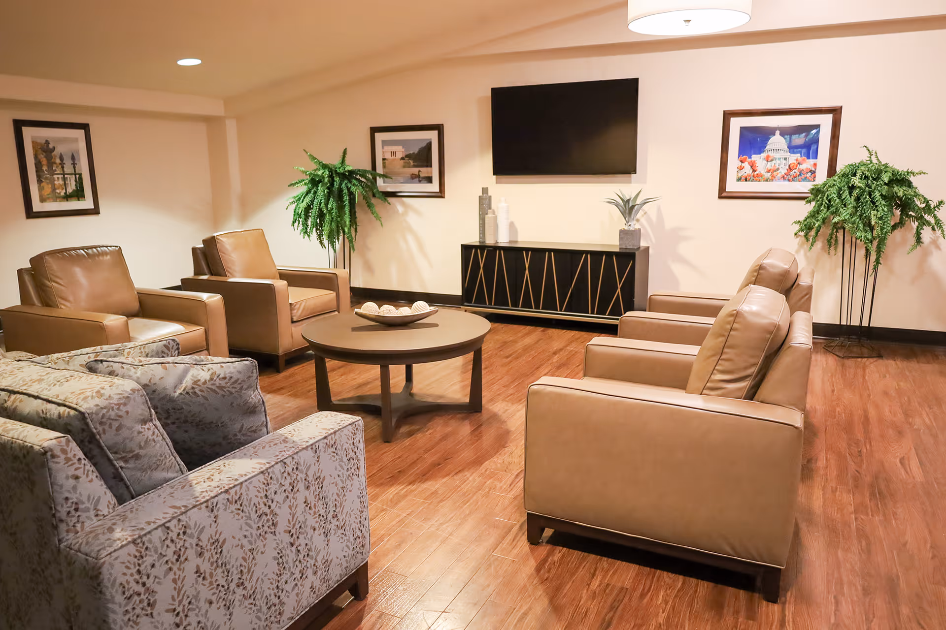 A cozy living room area with four brown leather armchairs and one patterned fabric armchair arranged around a round wooden coffee table. The room has wooden flooring, two green potted plants, framed pictures on the walls, and a flat-screen TV mounted above a black and gold cabinet.