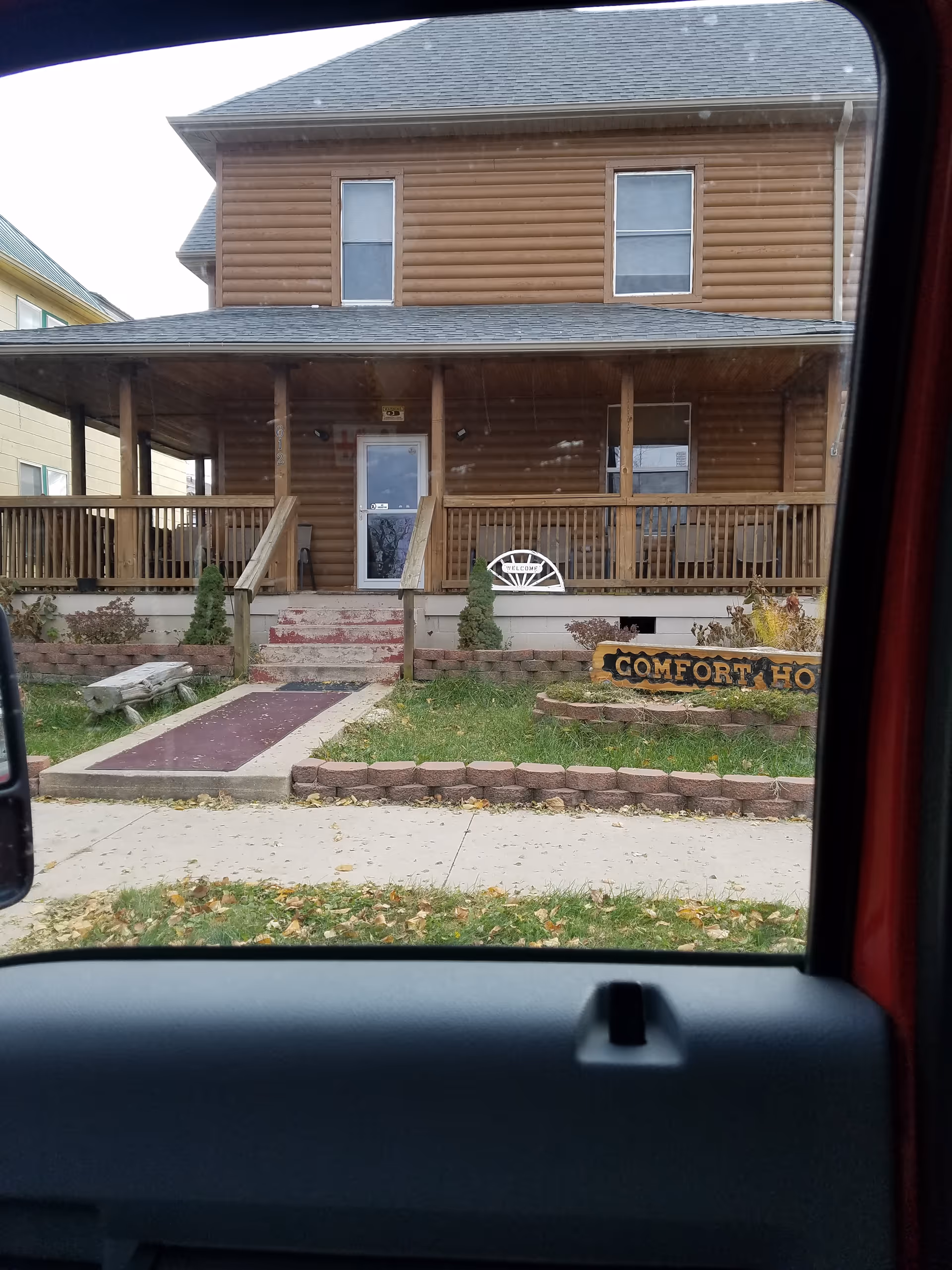 View of a two-story wooden house with a covered front porch, a ramp and stairs leading to the entrance, and a garden area with a sign that reads 'COMFORT HO'. The photo is taken from inside a vehicle, looking out through the window.