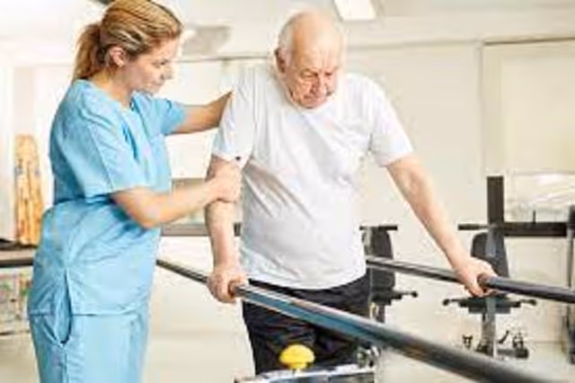 A healthcare worker in blue scrubs assists an elderly man who is using parallel bars for walking practice in a rehabilitation setting.