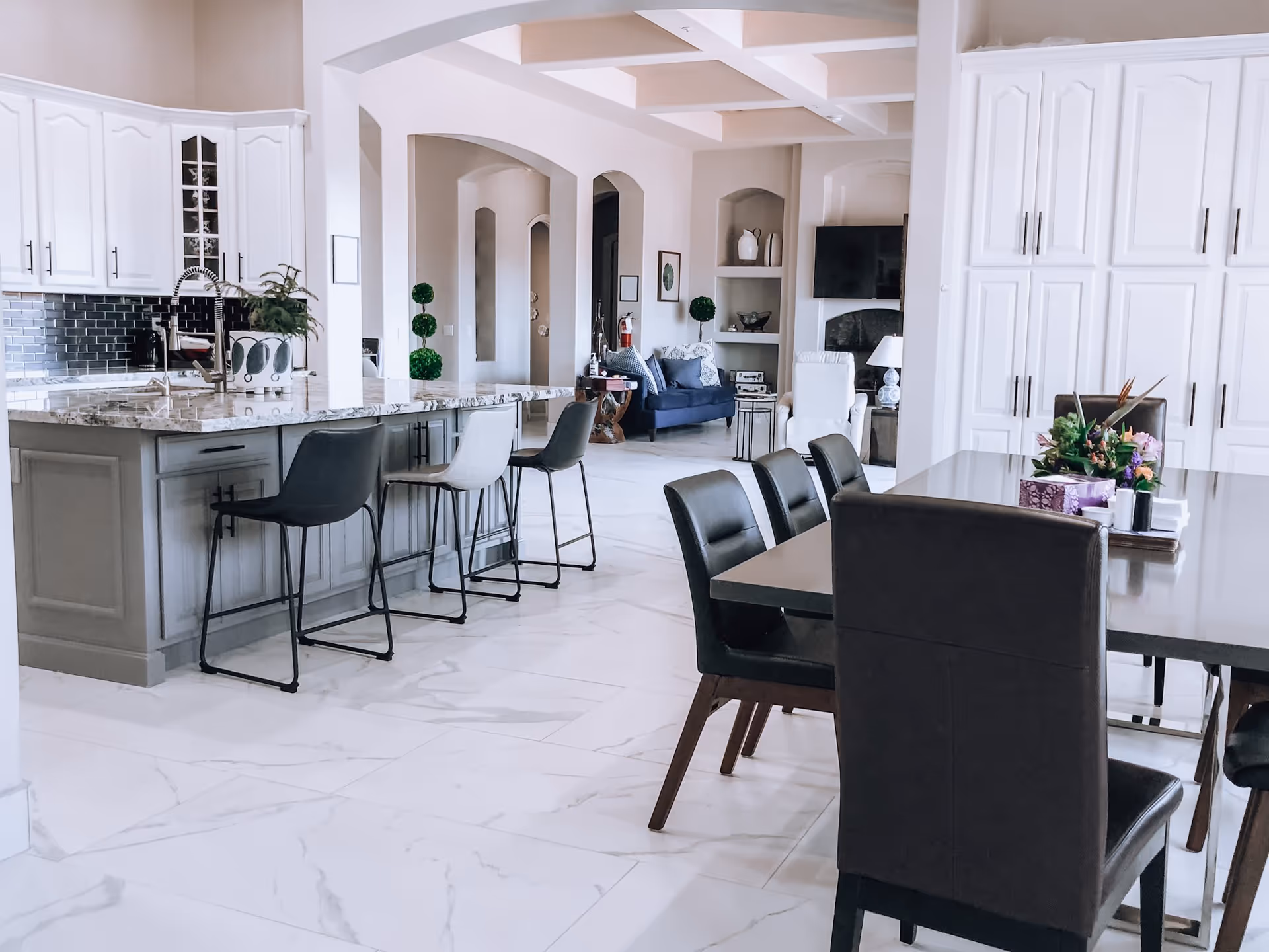 A bright, modern senior living facility interior featuring a kitchen area with a marble countertop island and bar stools, adjacent to a dining table with dark chairs. The background shows a cozy living area with a blue sofa, white armchair, and a wall-mounted TV. The space has white cabinetry, light-colored walls, and large marble floor tiles.