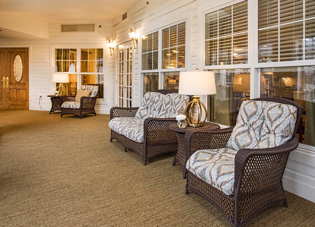 Wicker cushioned chairs and side tables with lamps arranged along a windowed interior lounge area.