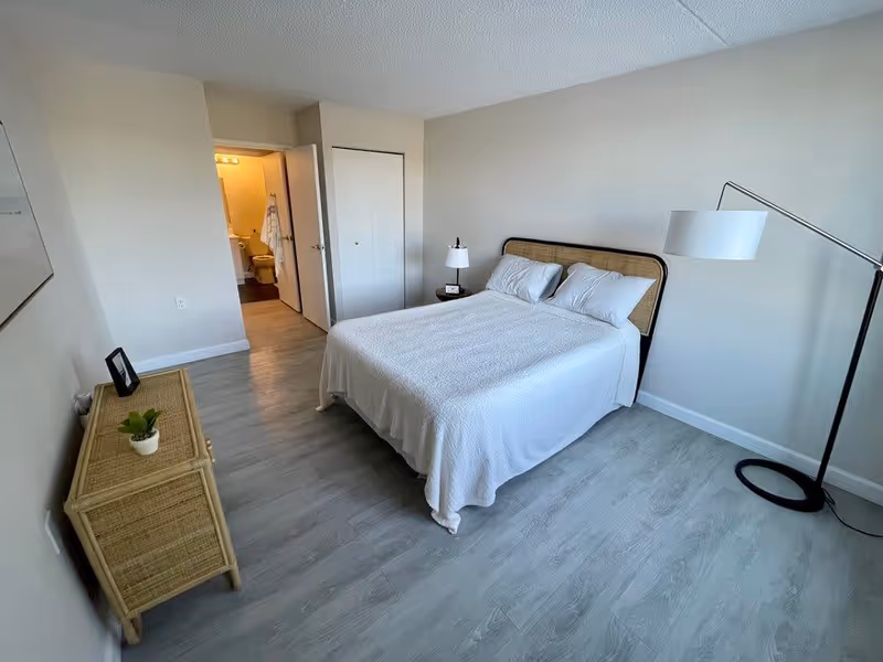 A bedroom with a neatly made bed covered in a white bedspread, two pillows, a bedside table with a lamp and clock, a floor lamp, a wicker dresser with a small plant and picture frame, light wood flooring, and a door leading to a bathroom.