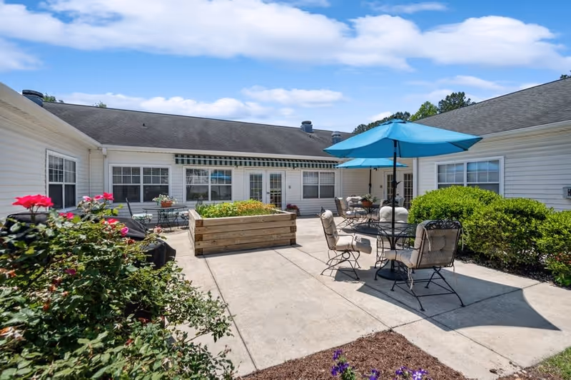 Outdoor patio area at Home Place of New Bern featuring a concrete courtyard with metal tables and cushioned chairs, one table shaded by a blue umbrella. Raised wooden garden beds and green bushes surround the seating area under a partly cloudy blue sky.