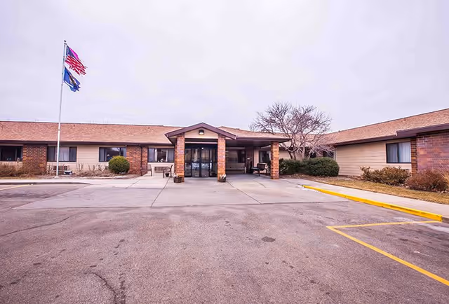 Front exterior view of Centennial Park Retirement Village building with a covered entrance, brick and beige siding, two flagpoles with American and state flags, and a parking area in front.
